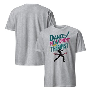 May include: Two light gray t-shirts. The front shirt has the words "DANCE MOVEMENT THERAPIST" in teal, pink, and yellow, with a black silhouette of a person dancing. The back of the shirt is plain.