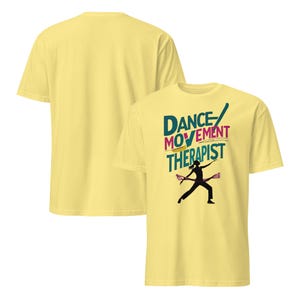 May include: Light yellow t-shirts with a crew neck. One shirt displays the text "DANCE MOVEMENT THERAPIST" in teal, pink, and yellow, with a black silhouette of a dancer. The other shirt is plain.