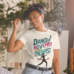 May include: A white t-shirt with the words "DANCE/MOVEMENT THERAPIST" in teal, pink, and yellow. A silhouette of a dancer is below the text. The person in the image is wearing the t-shirt and smiling.