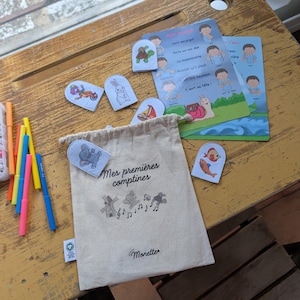 Peut inclure: Un sac en toile beige avec le texte "Mes premières comptines" et des illustrations de moulin à vent, de notes de musique et d'un éléphant dansant. Le sac est entouré de marionnettes à doigts en feutre et de cartes illustrées. Des feutres colorés sont également visibles.