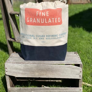 May include: A white canvas bag with a red and white label that reads "FINE GRANULATED THE NATIONAL SUGAR REFINING COMPANY NEW YORK, N.Y. AND PHILADELPHIA, PA". The bag is sitting on a wooden crate.