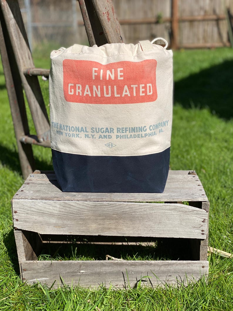 May include: A white canvas bag with a red and white label that reads "FINE GRANULATED THE NATIONAL SUGAR REFINING COMPANY NEW YORK, N.Y. AND PHILADELPHIA, PA". The bag is sitting on a wooden crate.