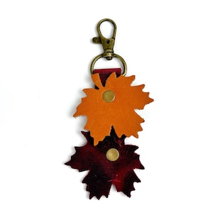 May include: A leather keychain featuring two maple leaf shapes in shades of orange and burgundy. The leaves are attached with brass rivets and a brass-colored clasp. The keychain is designed to be a decorative accessory.