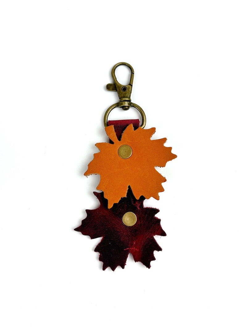 May include: A leather keychain featuring two maple leaf shapes in shades of orange and burgundy. The leaves are attached with brass rivets and a brass-colored clasp. The keychain is designed to be a decorative accessory.
