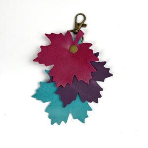 May include: A leather keychain featuring three leaf-shaped charms in shades of teal, purple, and burgundy. The charms are attached to a bronze-colored clasp. The leaves are layered and secured with a small, round rivet.
