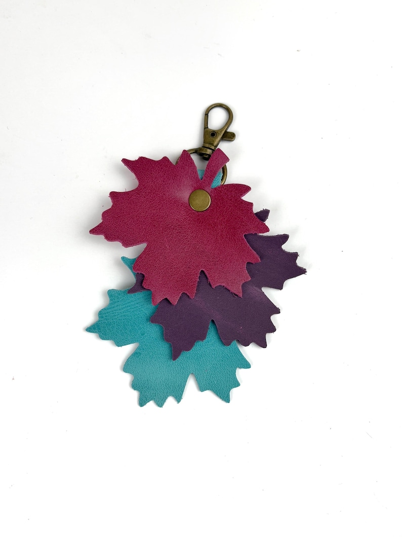 May include: A leather keychain featuring three leaf-shaped charms in shades of teal, purple, and burgundy. The charms are attached to a bronze-colored clasp. The leaves are layered and secured with a small, round rivet.