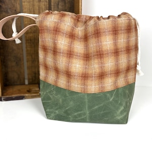 May include: A drawstring bag with a plaid top and a green bottom. The plaid pattern features shades of brown and tan. The bottom is a solid green color. The bag has a white drawstring and a leather handle.