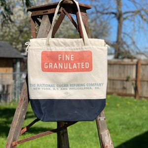 May include: A white canvas tote bag with a red and blue label that reads "FINE GRANULATED THE NATIONAL SUGAR REFINING COMPANY NEW YORK, N.Y. AND PHILADELPHIA, PA."