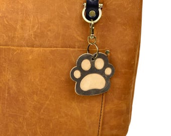 May include: A brown leather tote bag with a zipper closure and a black leather strap. A brass key chain with a paw print charm is attached to the strap.