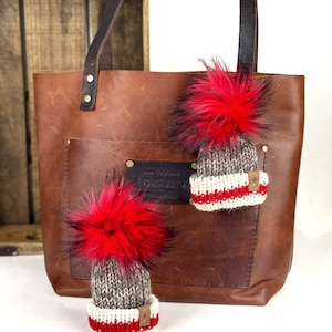 May include: A brown leather tote bag with a front pocket and dark brown handles. Two miniature knit hats with red fur pom-poms are displayed in front of the bag. The hats have a gray, white, and red color scheme.