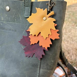 May include: A leather leaf keychain in shades of yellow, orange, and burgundy is attached to a green leather tote bag. The bag has a metal clasp and a button closure. The keychain is a fall-themed accessory.