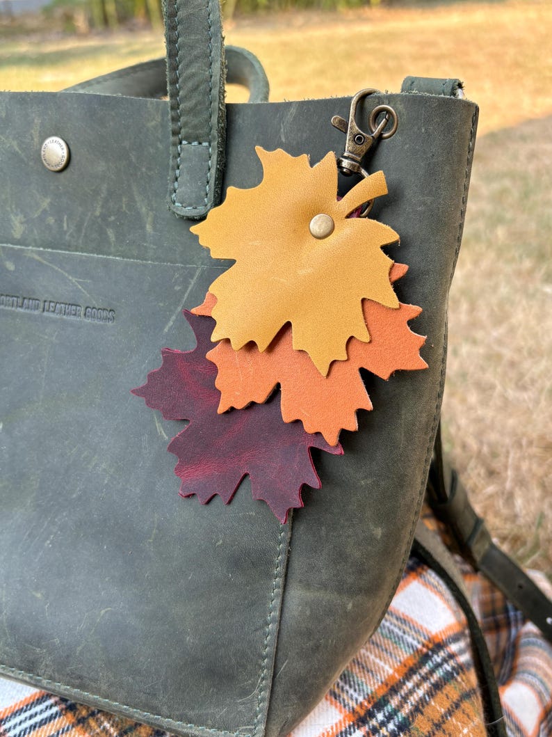 May include: A leather leaf keychain in shades of yellow, orange, and burgundy is attached to a green leather tote bag. The bag has a metal clasp and a button closure. The keychain is a fall-themed accessory.