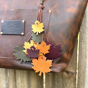 Könnte beinhalten: Eine braune Ledertasche mit einem dekorativen Blattanhänger. Der Anhänger zeigt Lederblätter in Orange, Gelb, Grün und Burgunderrot. Die Tasche hat einen Reißverschluss und ein kleines Lederetikett mit der Aufschrift "PORTLAND LEATHER GOODS".