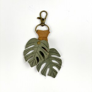 May include: A green leather keychain with two monstera leaf charms attached to a brown leather strap and a gold metal clasp.