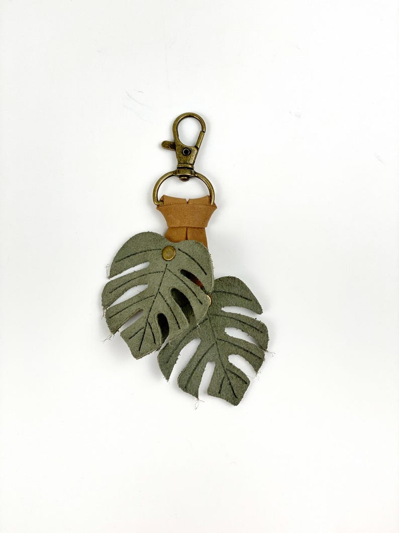 May include: A green leather keychain with two monstera leaf charms attached to a brown leather strap and a gold metal clasp.