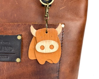 May include: A brown leather bag charm shaped like a Highland cow with a light-colored wooden muzzle and horns. The charm is attached to a brass-colored metal lobster clasp and ring, which is clipped to a dark brown leather strap on a large brown leather tote bag.