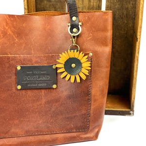 May include: A brown leather tote bag with a sunflower leather charm attached to the handle. The bag has a pocket on the front with a leather patch that reads "Hand Made Portland Leather Goods".
