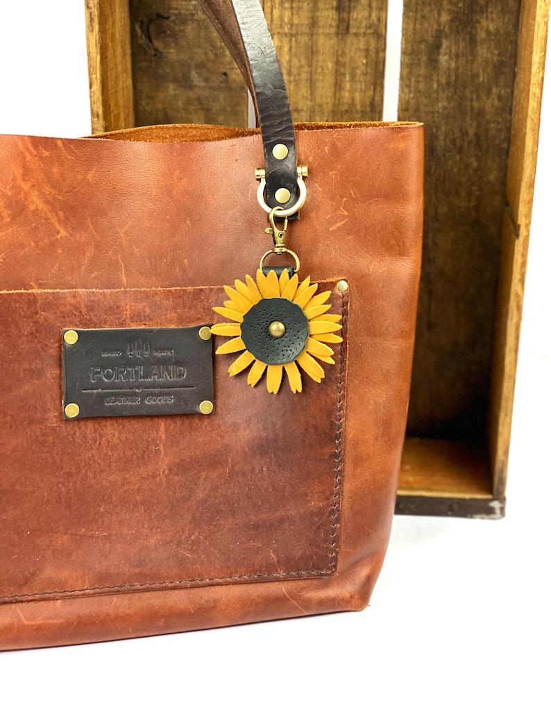 May include: A brown leather tote bag with a sunflower leather charm attached to the handle. The bag has a pocket on the front with a leather patch that reads "Hand Made Portland Leather Goods".