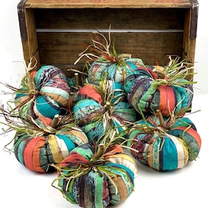 May include: A selection of fabric pumpkins in a variety of colours, including teal, orange, and brown, with vertical stripes. Each pumpkin has a twig stem and dried grass. They are displayed in front of a wooden crate.
