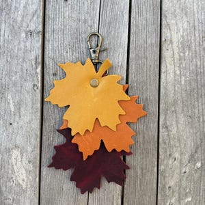 May include: A leather keychain featuring three layered maple leaf shapes in shades of yellow, orange, and burgundy. The keychain has a brass-colored clasp and a small button detail. The background is a weathered wooden surface.