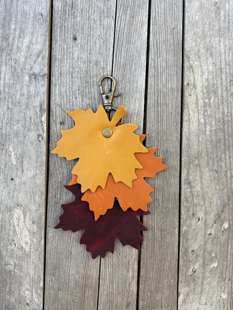 May include: A leather keychain featuring three layered maple leaf shapes in shades of yellow, orange, and burgundy. The keychain has a brass-colored clasp and a small button detail. The background is a weathered wooden surface.