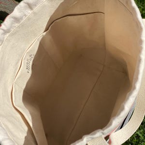 May include: A white canvas tote bag with a drawstring closure and a single strap. The bag has a small pocket on the inside and the text "WAY & THING" is printed on the inside of the bag.