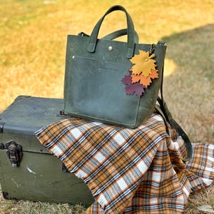 Puede incluir: Un bolso tote de cuero verde oliva con asa superior y bandolera desmontable. El bolso tiene un pequeño bolsillo delantero y un charm decorativo de hojas. Descansa sobre un baúl vintage cubierto con una manta a cuadros.