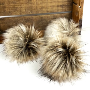 May include: Two fluffy brown and white faux fur pom poms. The pom poms are soft and fluffy and would be perfect for adding a touch of whimsy to a hat or scarf.