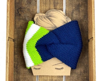 May include: A knitted headband featuring a twisted front design with color-blocked stripes in navy blue, lime green, and white. The headband is displayed on a tan cardboard cutout shaped like a head with closed eyes and a stylized hair bun.