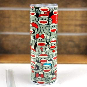May include: A stainless steel tumbler with a white background and a pattern of gray monkeys wearing red and blue hats. The monkeys are wearing red bows and some have red hearts on their chests.