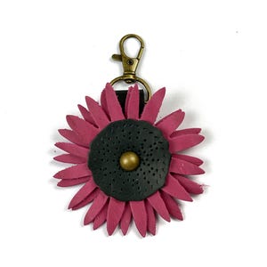 May include: A pink leather flower keychain with a black center and a gold button. The keychain has a brass clasp.