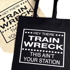 May include: Two tote bags with the text "Hey there... Train Wreck... This ain't your station" printed on them. One bag is black with white text and the other is beige with black text.