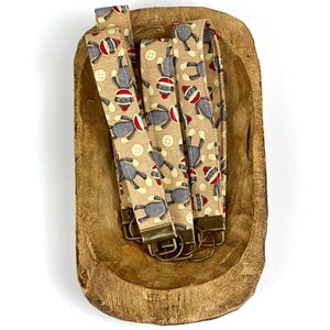 May include: Four tan fabric keychains with a pattern of monkeys wearing red and white hats. The keychains are in a wooden bowl and have gold key rings.