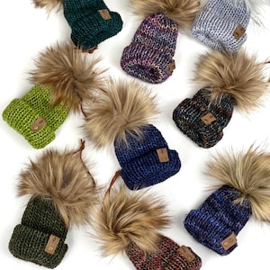 May include: Nine knitted hats with faux fur pom-poms. The hats are in various colors, including green, blue, purple, and gray. Each hat has a small leather tag with the word "Cozy" on it.