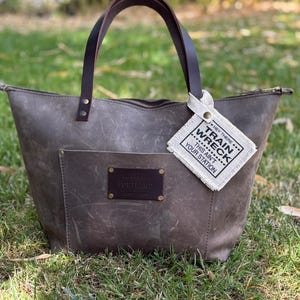 May include: A gray leather tote bag with dark brown leather handles, a front pocket, and a dark brown leather patch embossed with the word PORTLAND. A white fabric tag attached to the handle features the text HEY THERE TRAIN WRECK THIS AINT YOUR STATION.