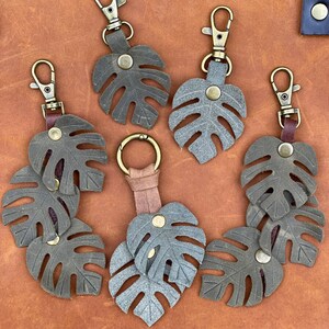May include: Six leather keychains with a monstera leaf design. The keychains are in shades of brown and green and have antique brass hardware.