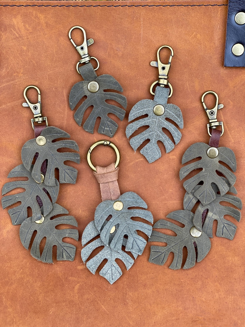 May include: Six leather keychains with a monstera leaf design. The keychains are in shades of brown and green and have antique brass hardware.