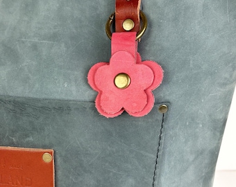 May include: A slate blue leather tote bag featuring a tan leather handle, a pink leather flower charm attached to a brass ring, and a rectangular tan leather patch with the embossed text "HAND MADE" and "GOODS" secured by brass rivets.