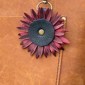 May include: A close-up of a leather bag with a burgundy leather flower charm attached to a key ring. The flower has a black center with a gold button.