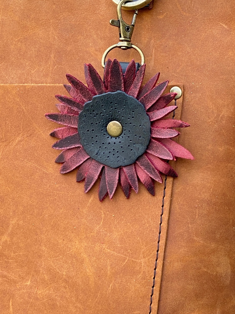 May include: A close-up of a leather bag with a burgundy leather flower charm attached to a key ring. The flower has a black center with a gold button.