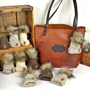 Puede incluir: Un bolso tote de cuero marrón con un bolsillo y una etiqueta de metal que dice "Old Portland". El bolso está rodeado de ocho gorros de punto con pompones de piel sintética en varios colores, incluyendo marrón, blanco y beige.
