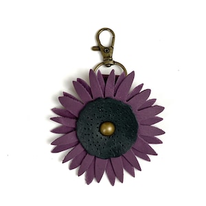 May include: A purple leather flower keychain with a black center and a gold button. The keychain has a metal clasp.