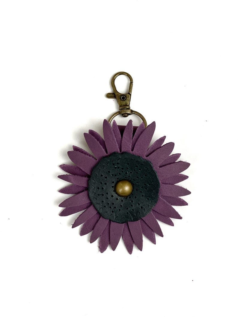 May include: A purple leather flower keychain with a black center and a gold button. The keychain has a metal clasp.