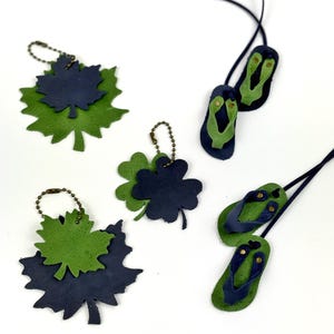 May include: Leather leaf and flip-flop pendants and keychains in green and navy blue. The leaf pendants include maple and four-leaf clover designs. The flip-flop pendants have green soles and navy blue straps. All items have a black cord or chain.