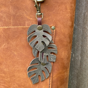 May include: A brown leather tote bag with a brass key chain featuring three layered, green metal monstera leaves.