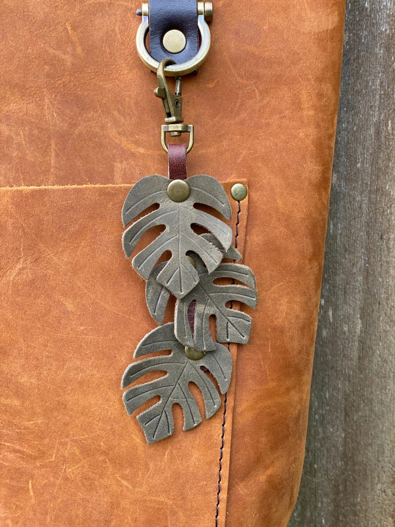 May include: A brown leather tote bag with a brass key chain featuring three layered, green metal monstera leaves.