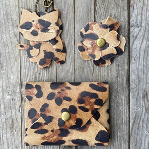 May include: A set of three small leather accessories in a leopard print pattern. The set includes a keychain with a cat charm, a flower charm, and a small wallet with a snap closure.