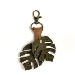 May include: A green leather keychain with two monstera leaf charms and a brown leather strap attached to a gold metal key ring.