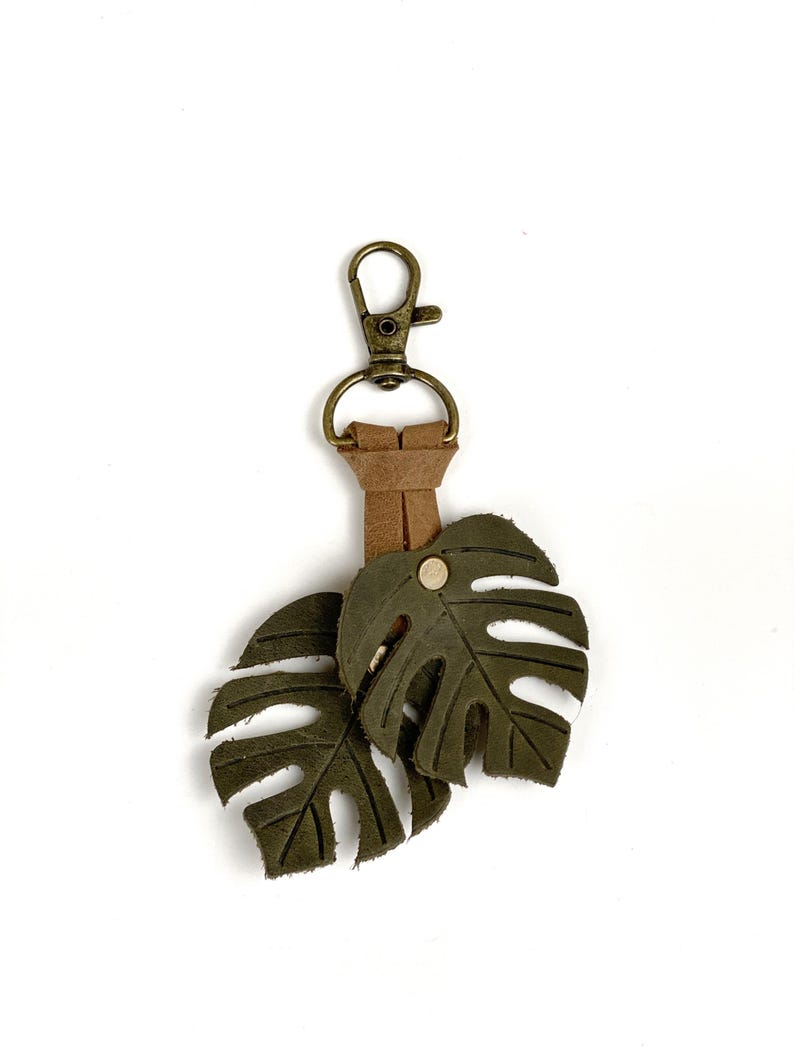 May include: A green leather keychain with two monstera leaf charms and a brown leather strap attached to a gold metal key ring.