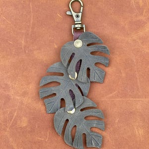 May include: A brown leather keychain with three dark gray leather monstera leaves attached to a brass key ring.